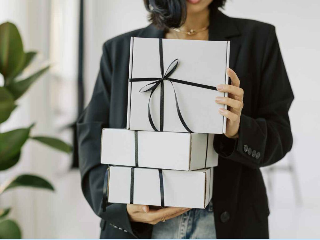 Imagem mostrando uma pessoa segurando três caixas de presente brancas empilhadas, todas amarradas com fitas pretas
