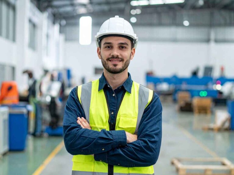 Engenheiro de segurança usando capacete branco e colete refletivo, de braços cruzados, em ambiente industrial com máquinas ao fundo.