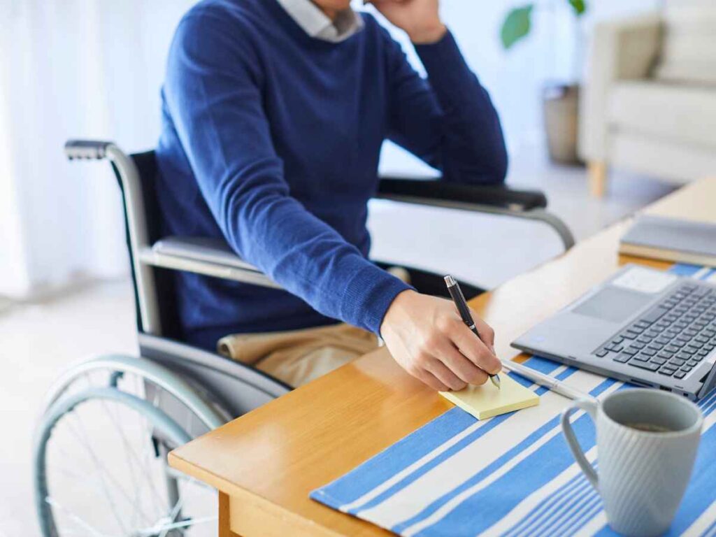 pessoa em cadeira de rodas escrevendo em bloco de notas enquanto trabalha em frente ao notebook
