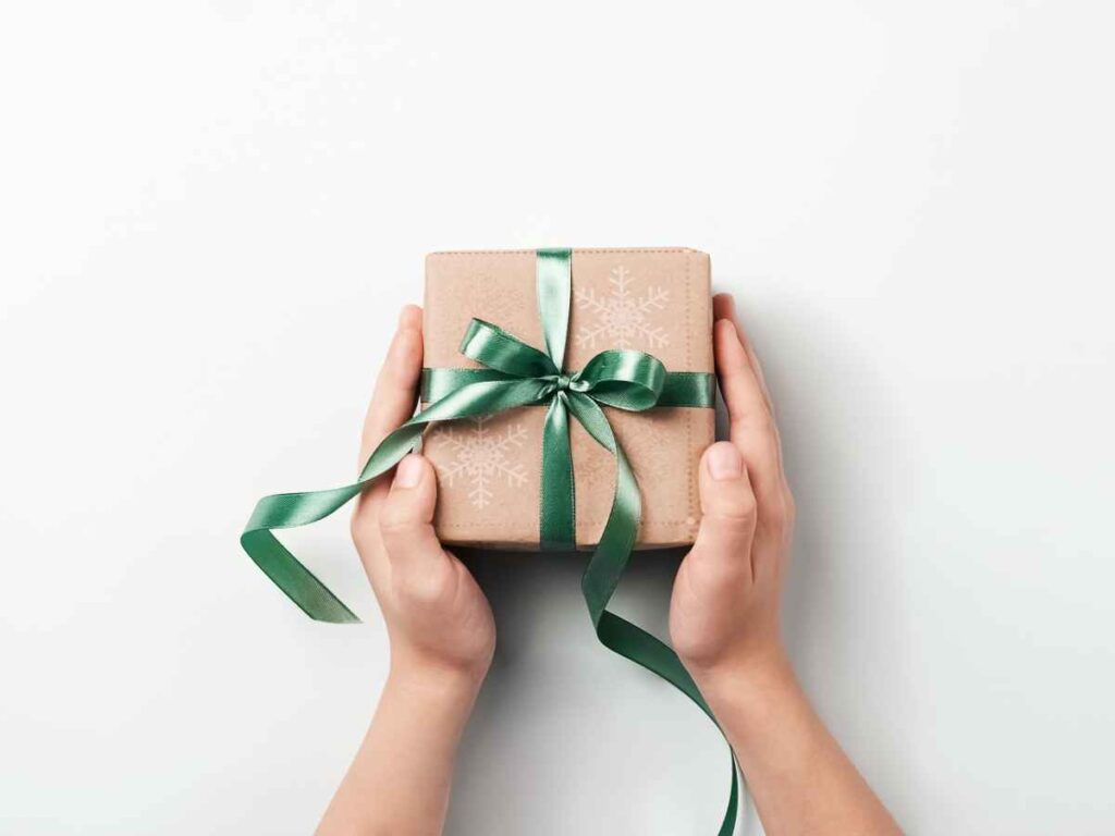 Mãos segurando uma caixa de presente embrulhada em papel kraft com laço verde brilhante, vista de cima