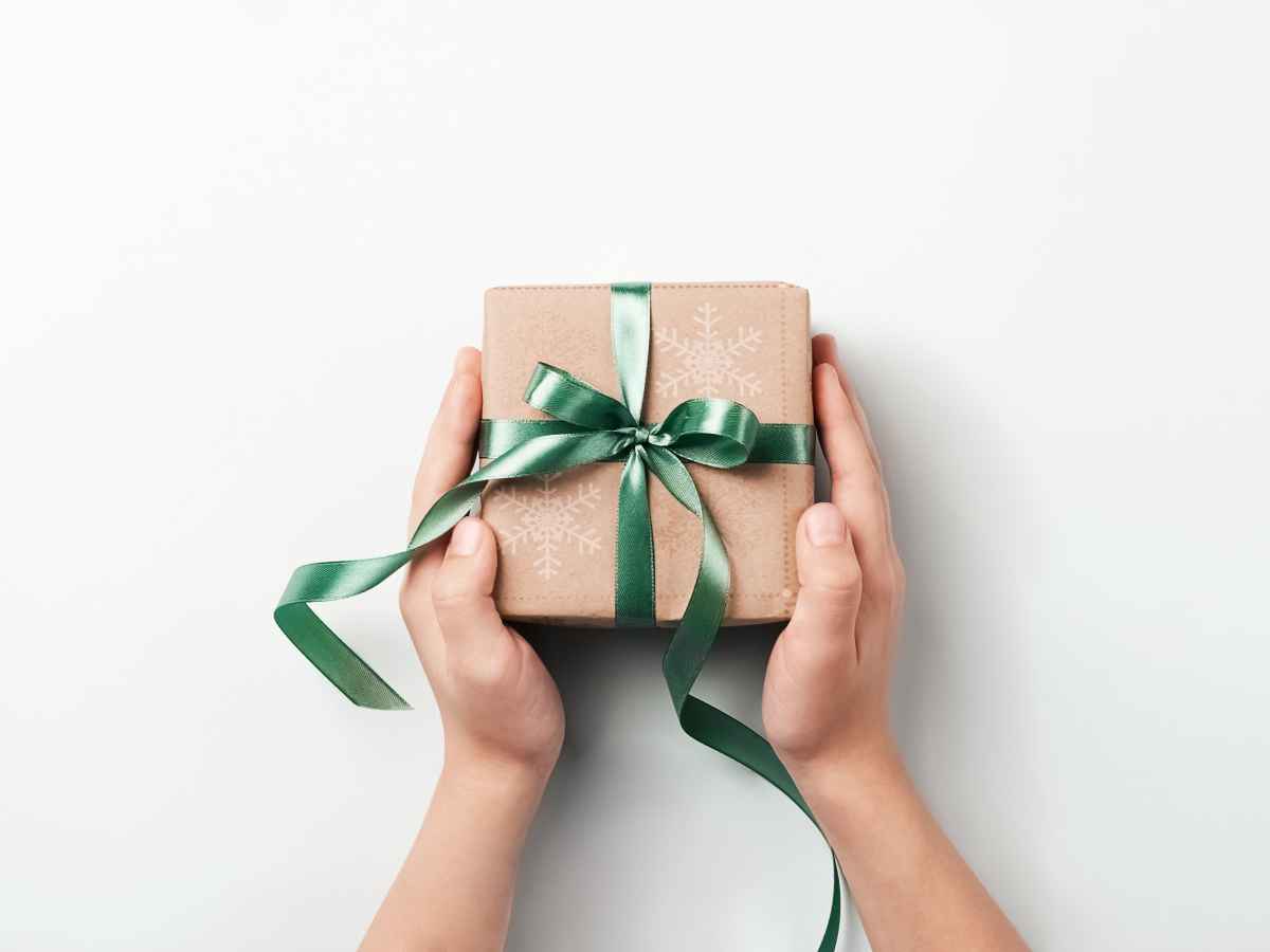 Mãos segurando uma caixa de presente embrulhada em papel kraft com laço verde brilhante, vista de cima