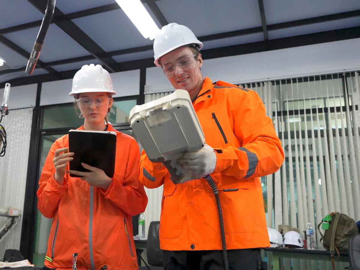 Dois técnicos de segurança usando capacetes e casacos de proteção laranja analisam equipamentos industriais