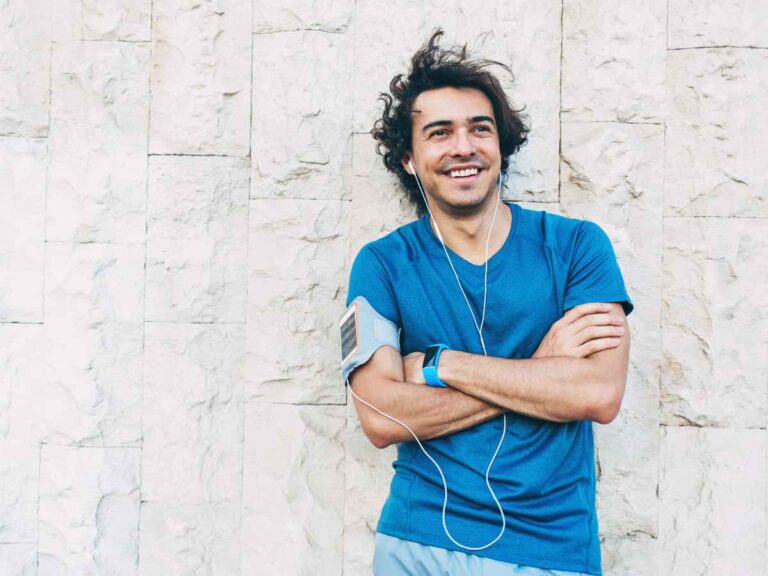 Homem sorridente usando fones de ouvido e roupa esportiva, encostado em uma parede