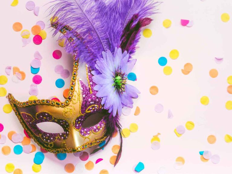 Máscara de carnaval colorida decorada com plumas roxas, flor e detalhes dourados, posicionada sobre fundo claro com confetes espalhados.
