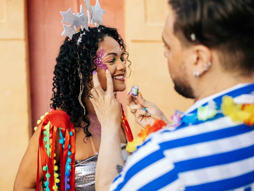 Pessoa aplicando maquiagem com glitter no rosto de mulher fantasiada para o carnaval