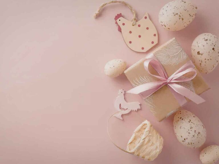 Presente de Páscoa embrulhado com fita rosa cercado por ovos decorativos e enfeites em formato de galinha sobre fundo rosa.