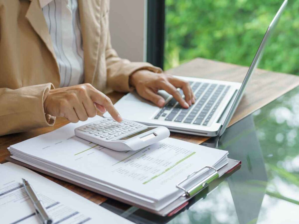 Pessoa usando calculadora enquanto trabalha no laptop com documentos sobre a mesa em ambiente de escritório. Simbolizando o planejamento de ações corporativas.